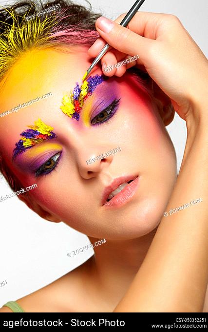 Female portrait with unusual face art makeup. Paint on brows, hair and around eyes. Artist's hand with paintbrush painting beautiful girl's brows make-up