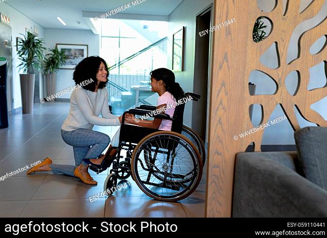 Happy mixed race mother kneeling holding hands talking with daughter in wheelchair in hospital foyer