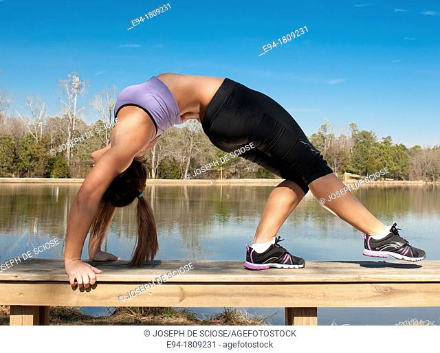 A 23 year old brunette woman in workout clothing doing a back bend on a bench by a pond