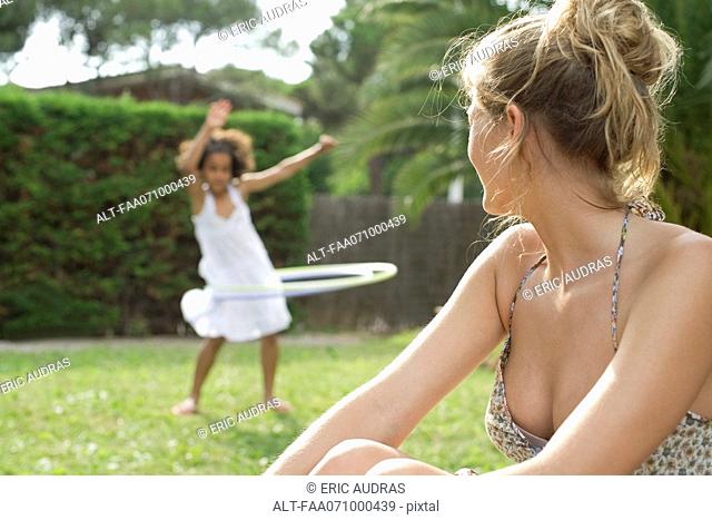 Woman sitting outdoors, watching child playing with plastic hoop