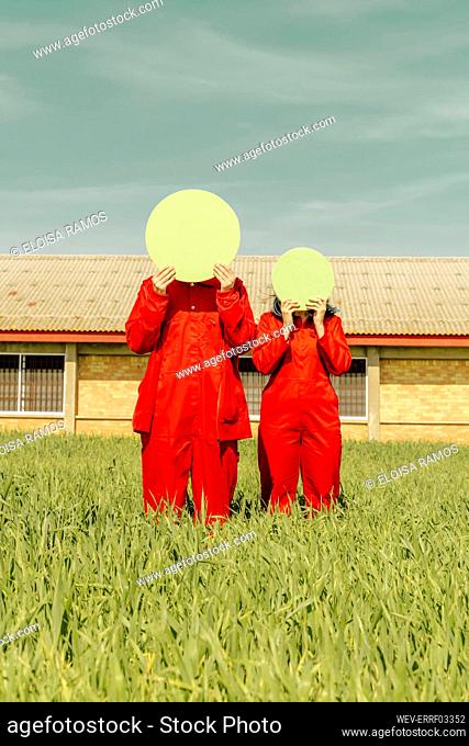 Young couple wearing red overalls standing on a field hiding faces behind green circle