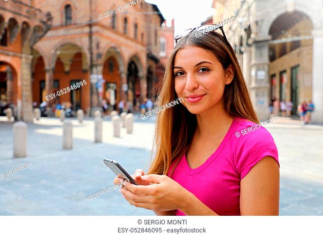 Happy woman smiling and using a smartphone in the street and looking at camera