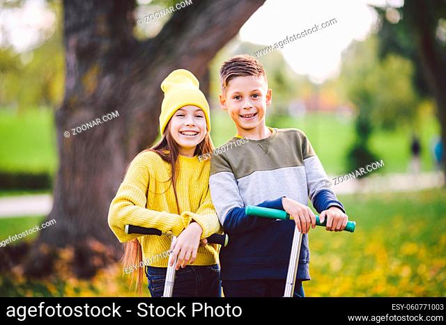 Caucasian children twins boy and girl schoolchildren age pose together in sunny weather in autumn park after riding on kick scooters