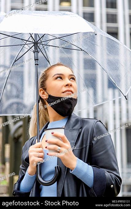 Thoughtful businesswoman wearing face mask holding umbrella and disposable cup in city during rainy season