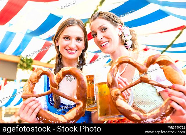 Two friends wearing dirndl are standing in a beer tent at Dult or Oktoberfest with giant pretzels