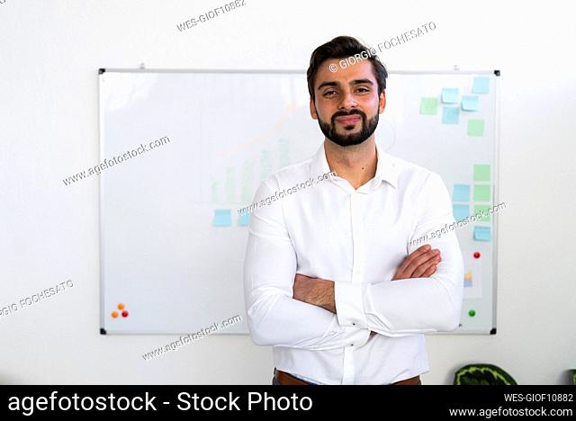 Smiling male entrepreneur with arms crossed standing against whiteboard in office