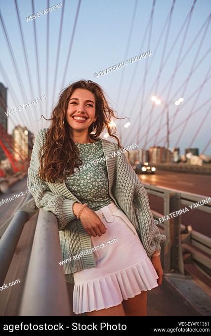 Smiling woman standing on bridge against sky in city during sunset