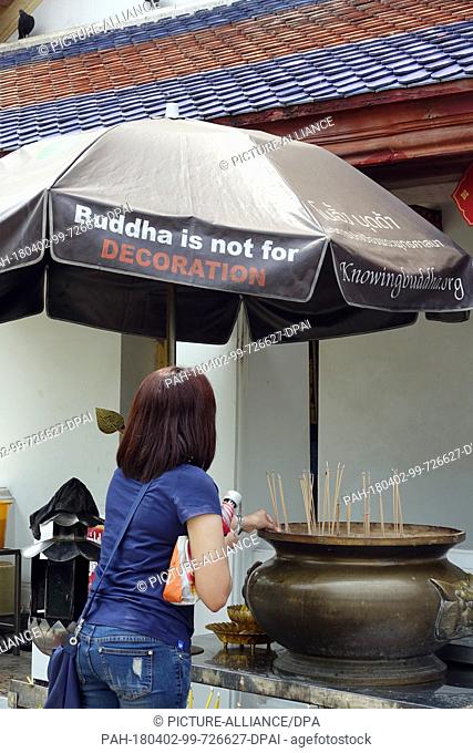 27 February 2018, Thailand, Bangkok: A woman prays at the Wat Arun (Temple of Dawn) and lights an incense. A sunshade reads 'Buddha is not for decoration'