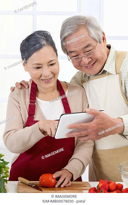 Senior couple holding touch pad in the kitchen and looking with smile together