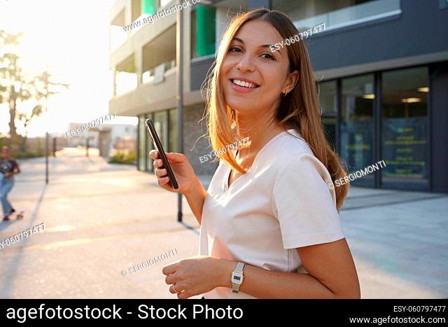 Dynamic confident businesswoman using mobile phone and looking at camera while standing on the street at sunset