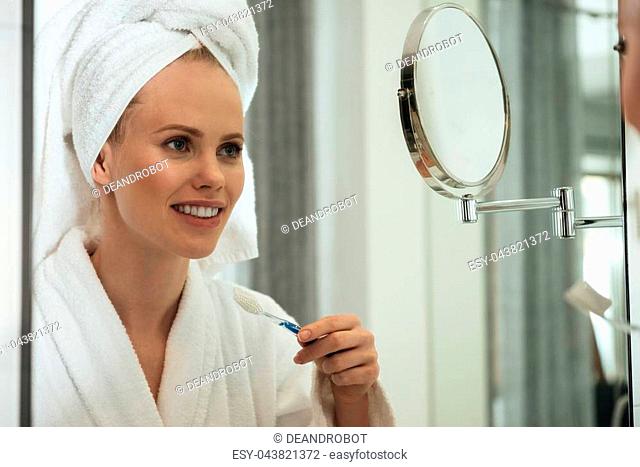 Young blonde woman with towel on head and wearing bathrobe brushing teeth against mirror in bathroom