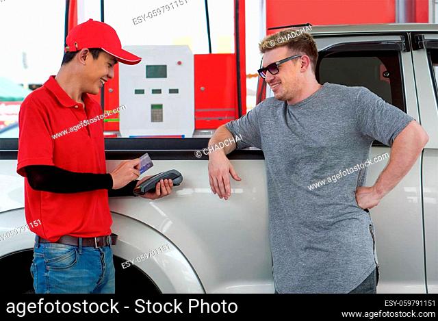 Gas station worker in red uniform stand smiling, swipe mockup credit card via payment terminal. Caucasian driver with sunglasses waiting beside bronze pickup...