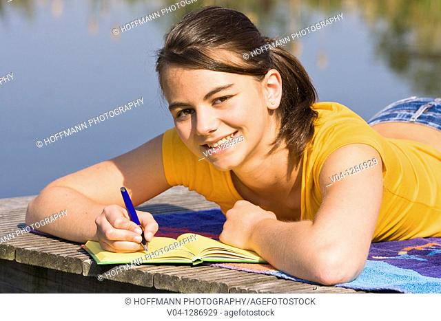 A young girl writing into her diary at a lake, smiling at the camera