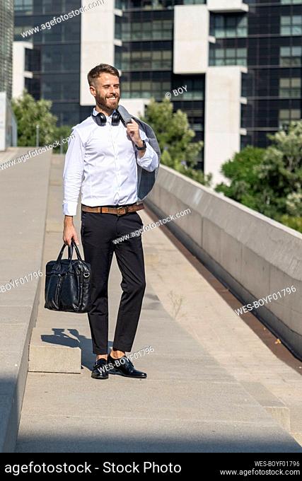 Businessman holding jacket and briefcase while standing on bridge during sunny day