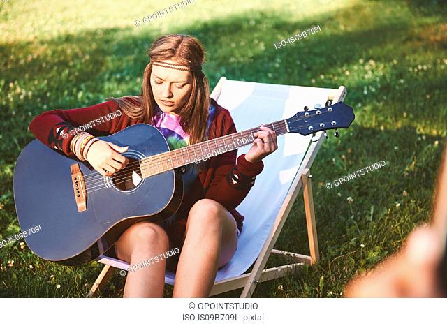 Young boho woman sitting on deckchair playing acoustic guitar at festival