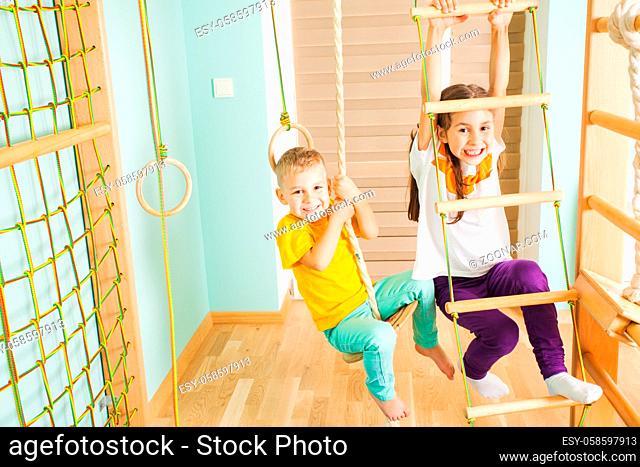 Happy smiling siblings boy and girl swinging on a wooden ladder and swing on sports complex. Close view of brother and sister happily playing on a kids gym in...