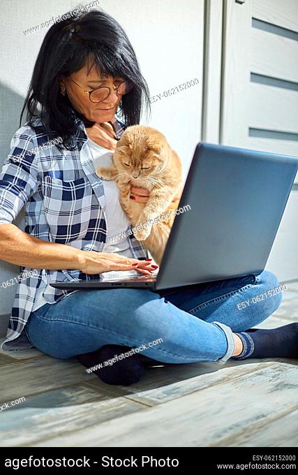 Mature Woman searching vet website on laptop to register cat for veterinary consultation, Freelancer female typing on laptop sitting on the floor at home