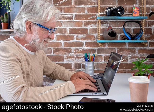 profile of elderly man browsing social media content using laptop on desk with disposable coffee cup. old retired man spending his time by working on laptop at...