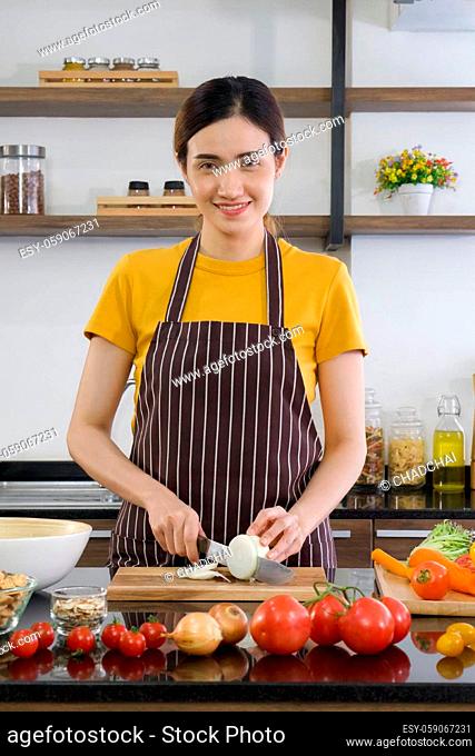 Young housewife slices onion into pieces on a wooden chopping board. Morning atmosphere in a modern kitchen