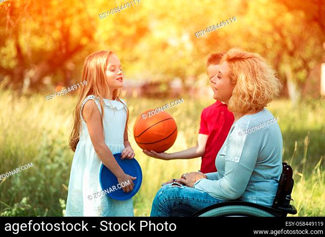 Beautiful Mother In A Wheelchair Talks With Her Laughing Kids. Small Girl In White Dress Holds Frisbee And Tells Some Story Her Smiling Mother In A Wheelchair...