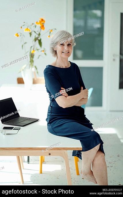 Smiling businesswoman with arms crossed sitting on desk in office