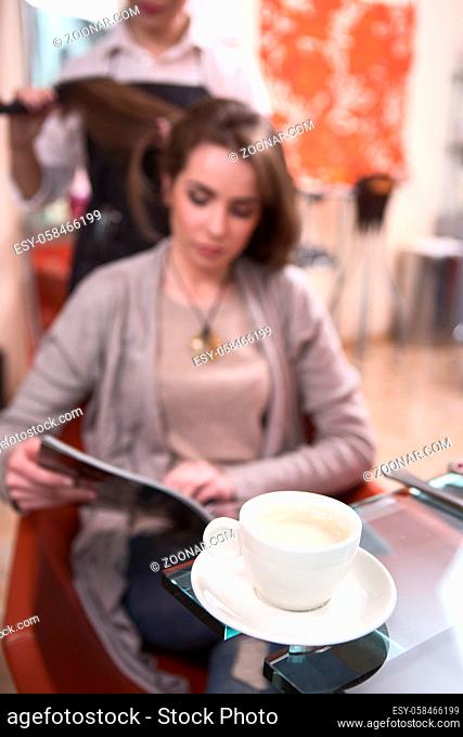Closeup picture of cup of coffee. Beautiful lady reading magazine while having her hair dobe by hairdresser in hairdressing saloon
