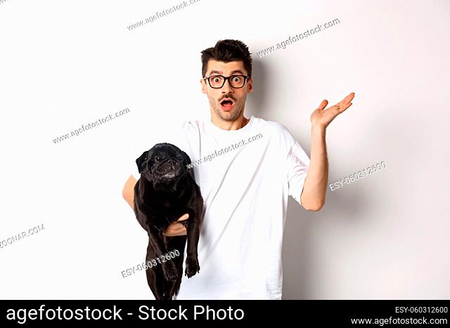 Image of confused hipster man holding dog and shrugging, dont know, raising hand puzzled, standing with his animal over white background