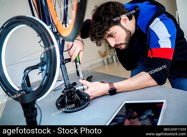 Concentrated young man fastening bicycle seat with wrench while filming at home