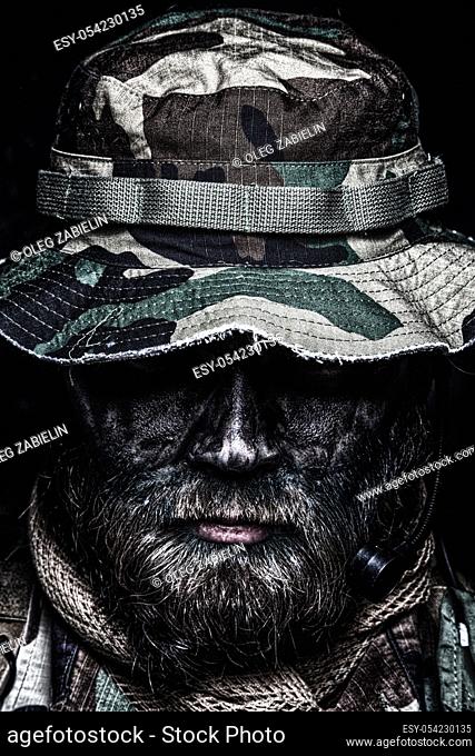 Close up, low key portrait of bearded commando fighter, army special forces soldier, private military company mercenary in boonie hat, tactical radio headset