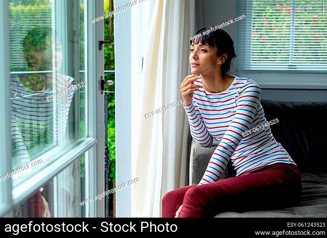 Thoughtful caucasian young woman looking through window while sitting at home