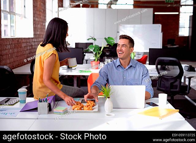 Happy caucasian male and female colleague eating pastries and talking sitting in office