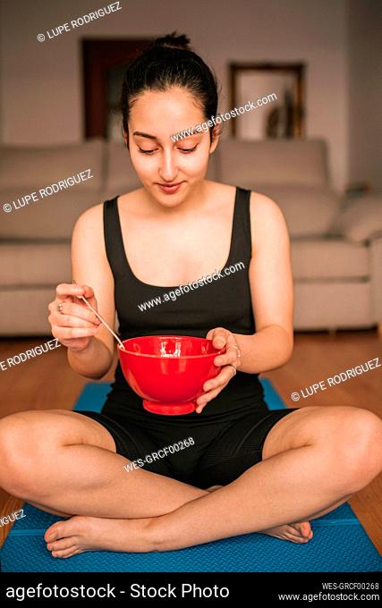 Young woman holding breakfast bowl while sitting on exercise mat at home