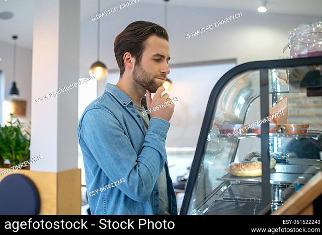 Difficulty of choice. Young bearded man standing sideways to camera touching his chin thoughtfully looking at food display in cafe