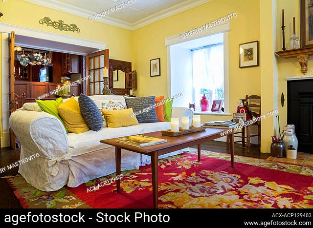 Teak wood coffee table, sofa with colourful cushions in living room with French doors leading into kitchen inside an old circa 1830 Quebecois style country home