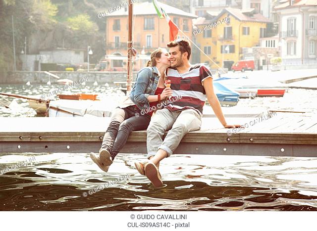 Couple sitting on pier whispering and eating ice cream cone at lake Mergozzo, Verbania, Piemonte, Italy