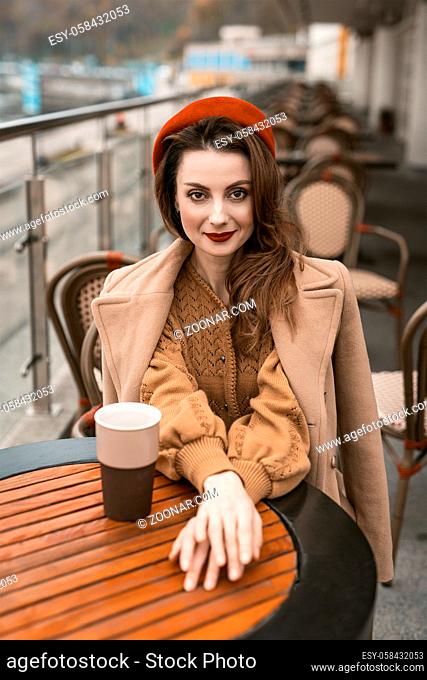 French woman drink coffee sitting at restaurant terrace with coffee mug looking at camera. Portrait of stylish young woman wearing autumn coat and red beret...