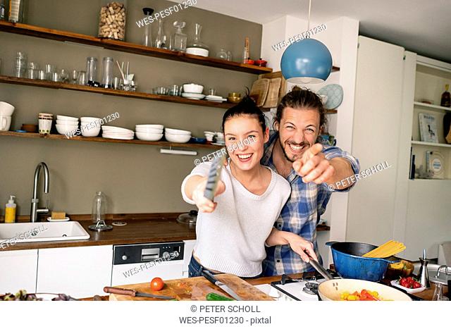 Young couple preparing food together, laughing and pointing with spatula