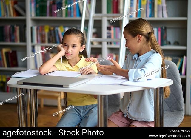 Smiling fair-haired adolescent girl seated at the library table tapping her bored friend on the shoulder