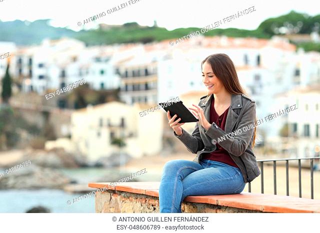 Happy woman using a tablet sitting on a ledge in a coast town on vacation