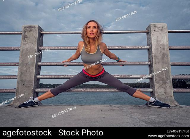 Young pretty blonde female wearing exercise clothing stretching on the pier near the sea and enjoying early morning with beautiful sunrise