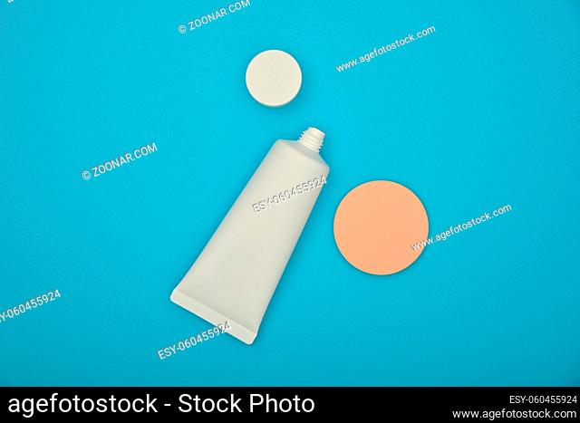 Close up one white tube of makeup foundation cream and beige sponge over blue background, elevated top view, directly above