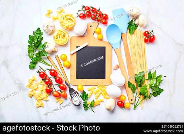 Small blackboard with copy space surrounded by ingredients for cooking different types of pasta, condiments, utensils, healthy raw vegetables scattered light...