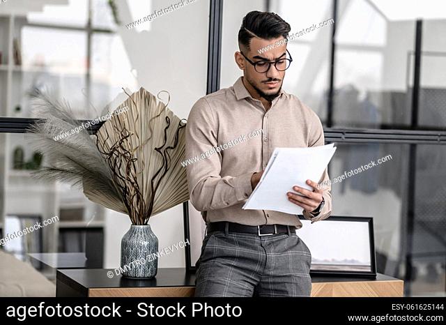 Work with documents. Well dressed young bearded man looking attentively at papers standing with back to glass wall at home