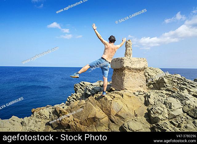 A man with a shirtless back and summer shorts leans on one foot and raises his breaches as a sign of victory and joy