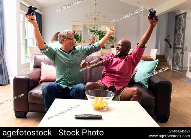 Multiracial senior male friends with arms raised enjoying video game at home