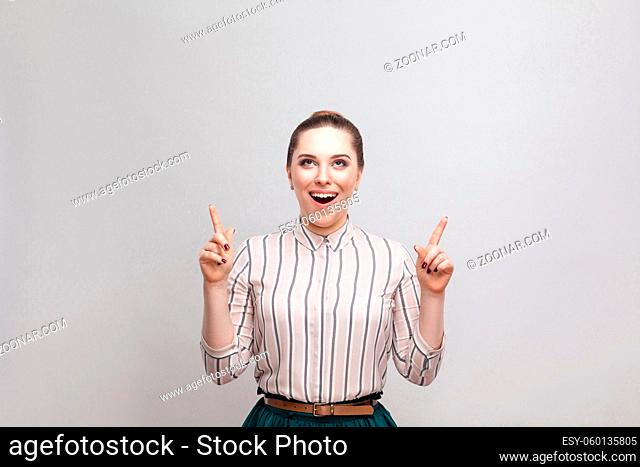Surprised beautiful young woman in striped shirt and green skirt with makeup and collected ban hairstyle, standing with amazed face and pointing at copyspace