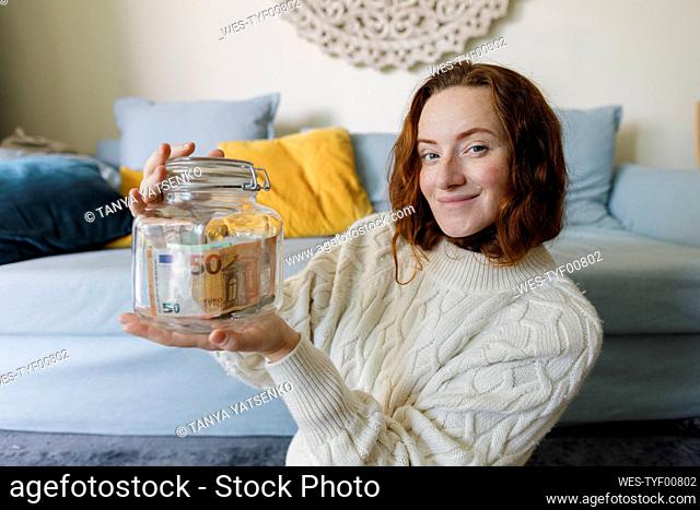 Smiling woman holding savings jar at home