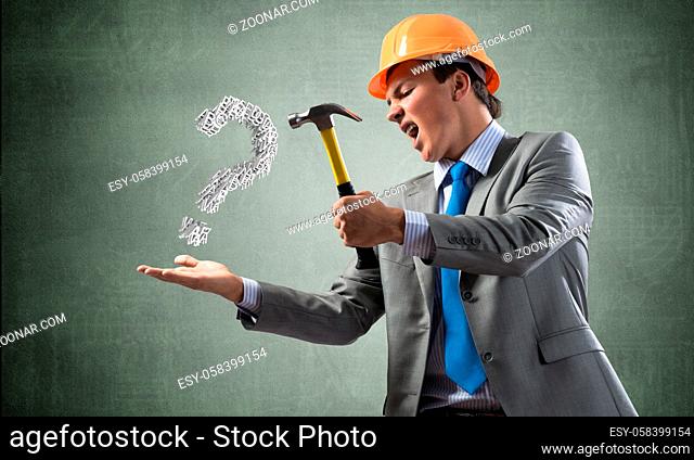 Furious businessman going to crash with hammer question mark. Young handsome man in business suit and safety helmet standing on wall background