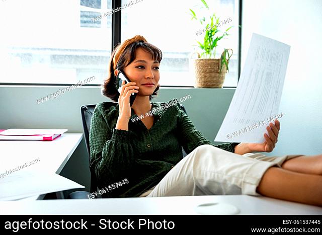 Young asian businesswoman examining document while talking on mobile phone in office