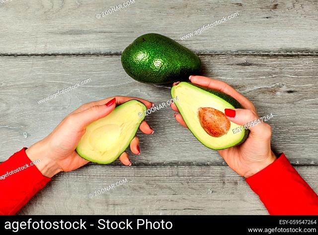 Tabletop view, woman hand holding two halves of avocado in each hand, one more whole pear placed on gray wooden desk above